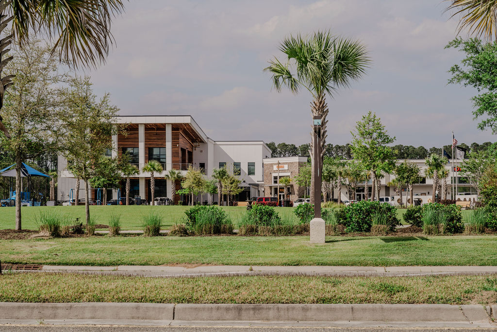 Apartments in Bluffton A modern building with large windows and a covered entrance sits behind a well-manicured lawn with small trees and shrubs. Palm trees are in the foreground with a road in the front.