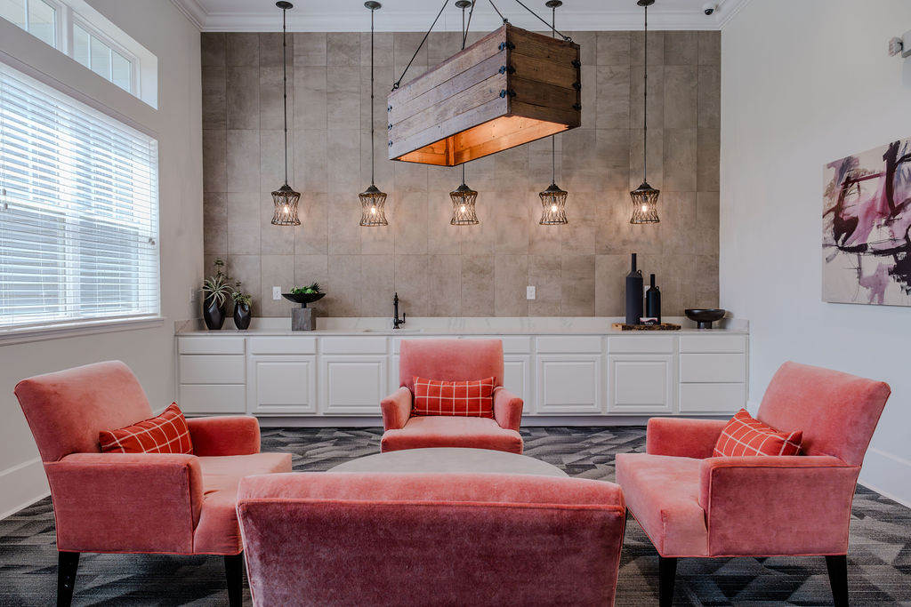 Apartments in Bluffton A modern living area with four pink chairs surrounding a round ottoman, under a wooden hanging light fixture. A white cabinet with a sink and decorative items is in the background.