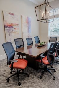 Apartments in Bluffton A modern conference room with a wooden table, adjustable office chairs, and abstract paintings on the wall. Natural light streams in through a window covered by horizontal blinds.