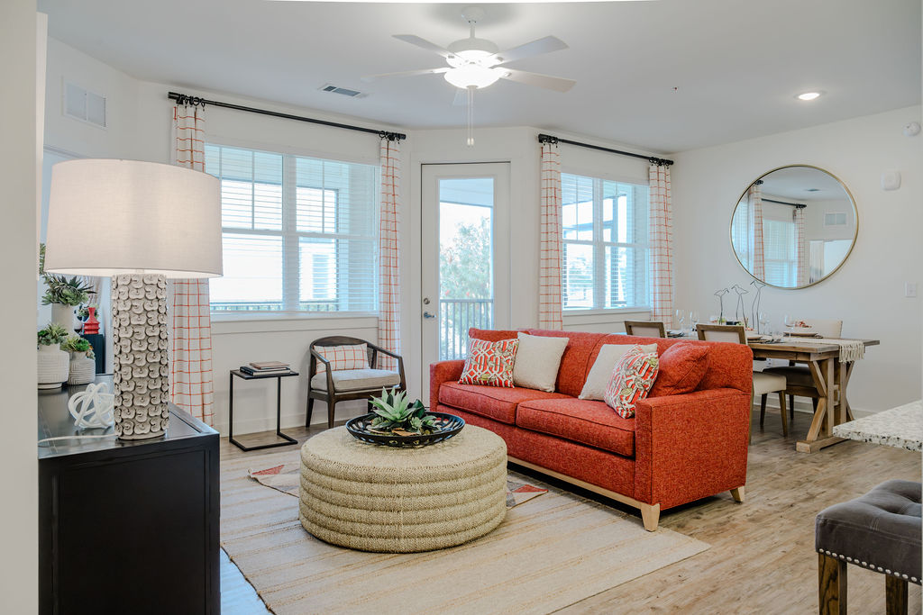 Apartments in Bluffton A modern living room with a red couch, a round woven coffee table, a lamp, a dining area with a wooden table, and a large round mirror on the wall. The room is well-lit with natural light.