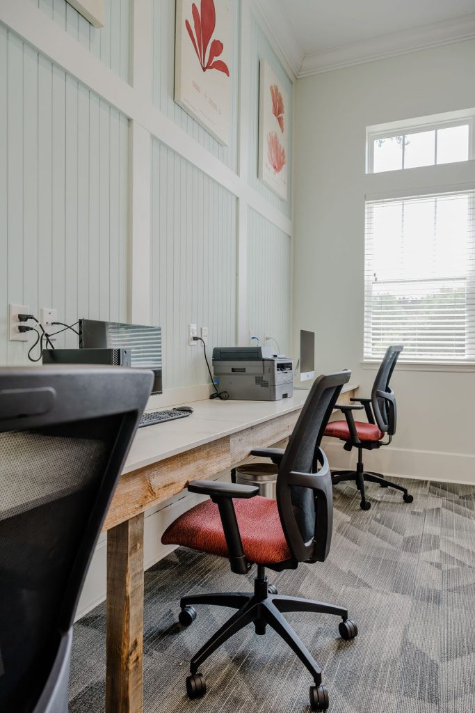 Apartments in Bluffton A modern office workspace with wooden desks, gray and red office chairs, computers, a printer, and decorative wall art. Bright room with natural light from large windows.