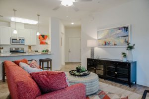 Apartments in Bluffton Spacious living room with a red sofa, round coffee table, and decorative plants. A modern kitchen with white cabinets is visible in the background. Wall art and a sideboard enhance the decor.