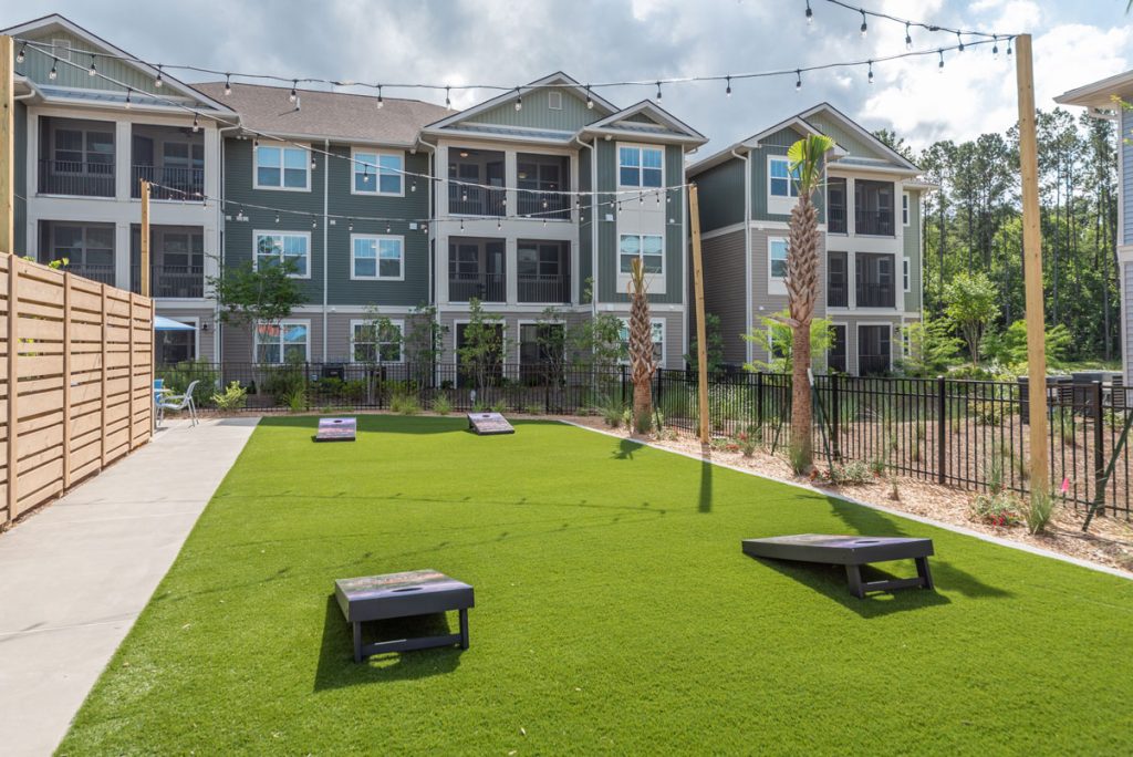 Apartments in Bluffton Outdoor recreation area with a neatly manicured lawn, cornhole boards, string lights overhead, and an apartment building in the background.