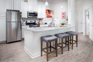 Apartments in Bluffton Modern kitchen with white cabinets, stainless steel appliances, large island with granite countertop, three gray cushioned stools, and colorful abstract art on the walls.