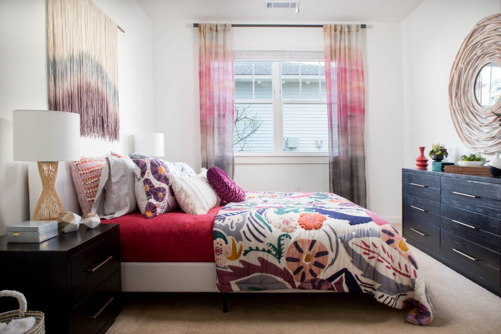 Apartments in Bluffton A bedroom interior featuring a bed with colorful floral bedding, red blanket, and multiple pillows. There's a window with ombre curtains, two dressers, bedside lamps, and various decorative items.