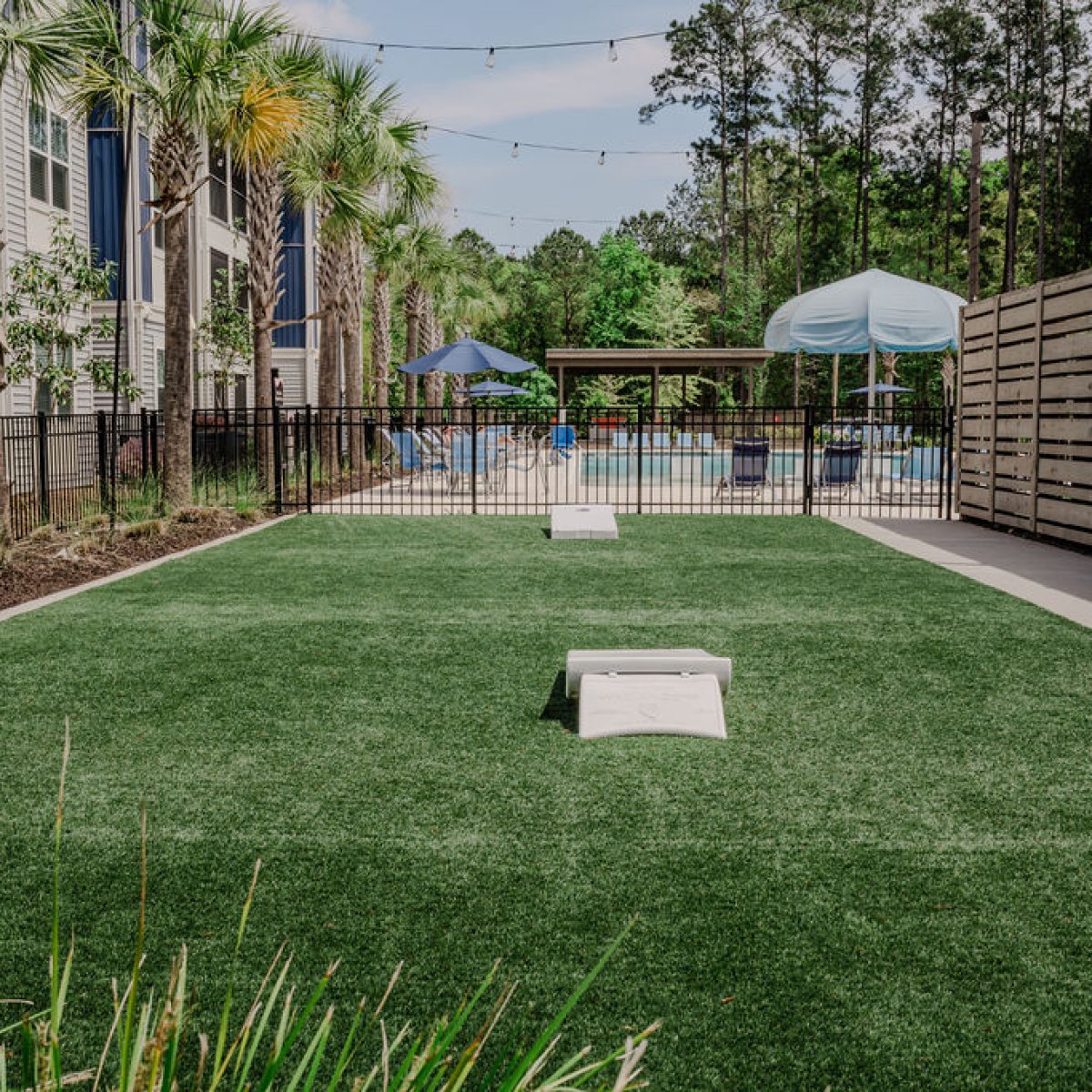 Apartments in Bluffton A fenced outdoor recreation area with artificial grass, two cornhole boards, a wooden privacy wall, and a pool surrounded by patio furniture and umbrellas in the background.