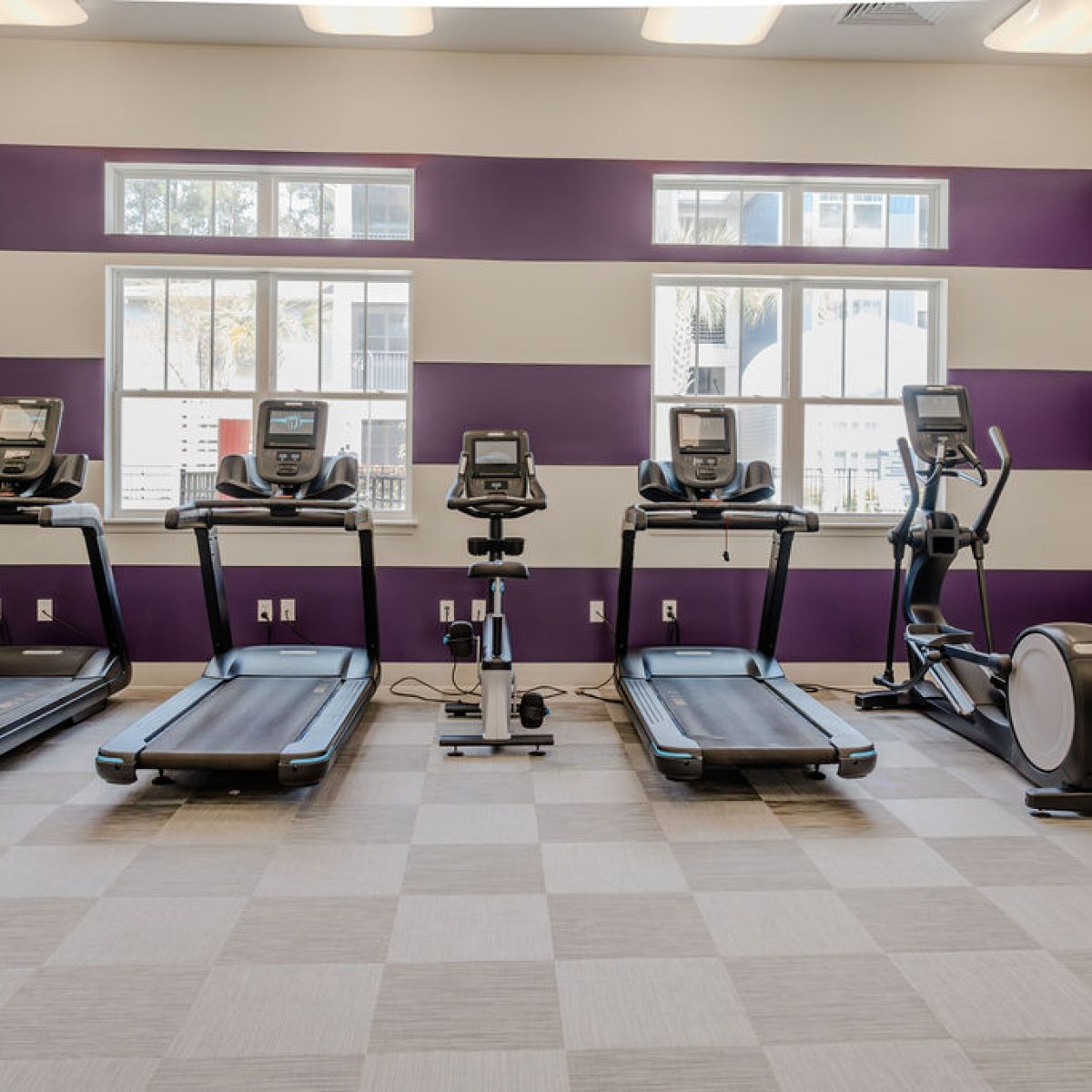 Apartments in Bluffton A modern gym with striped purple and white walls featuring three treadmills, two exercise bikes, and an elliptical machine, all facing windows that let in natural light.