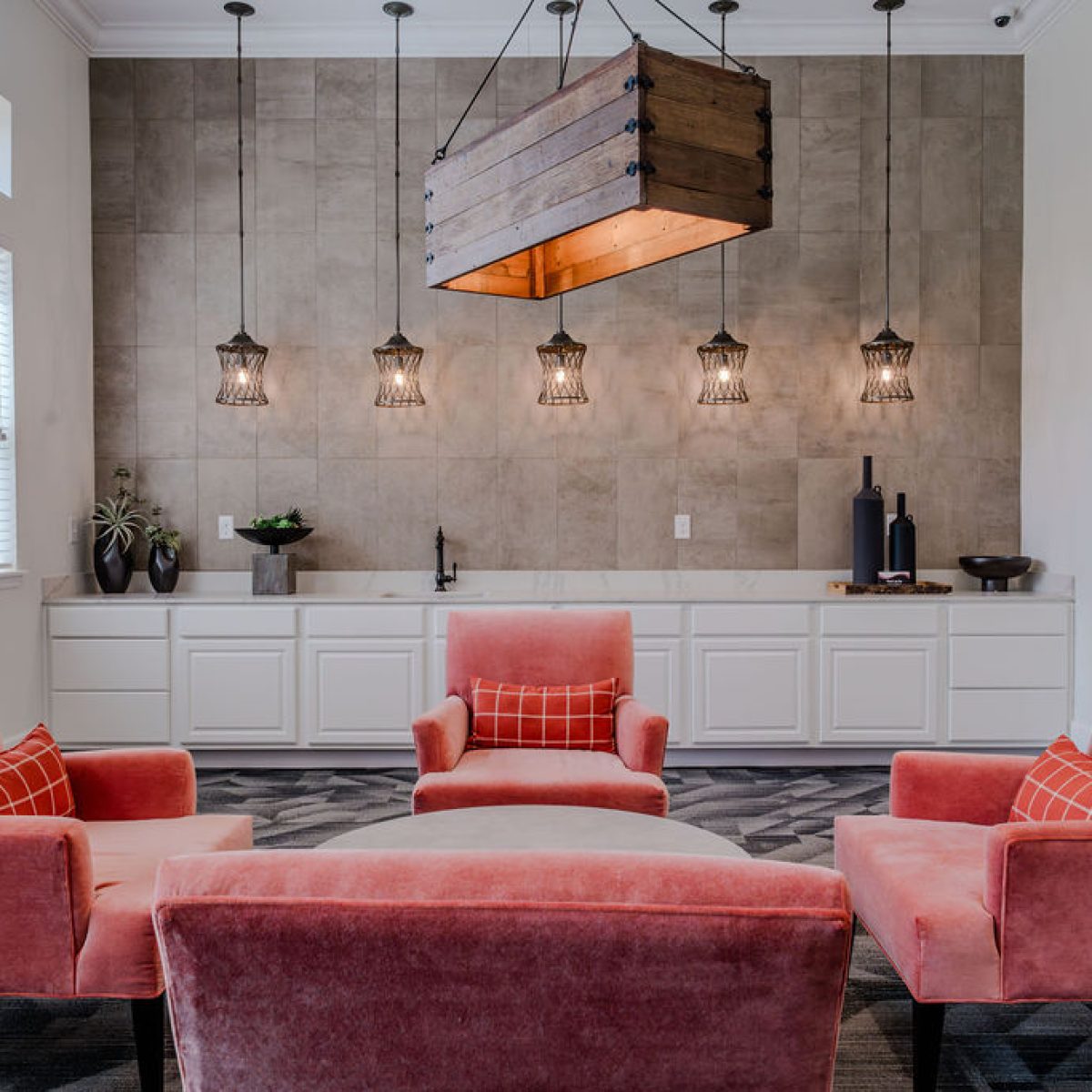 Apartments in Bluffton A modern living area with four pink chairs surrounding a round ottoman, under a wooden hanging light fixture. A white cabinet with a sink and decorative items is in the background.