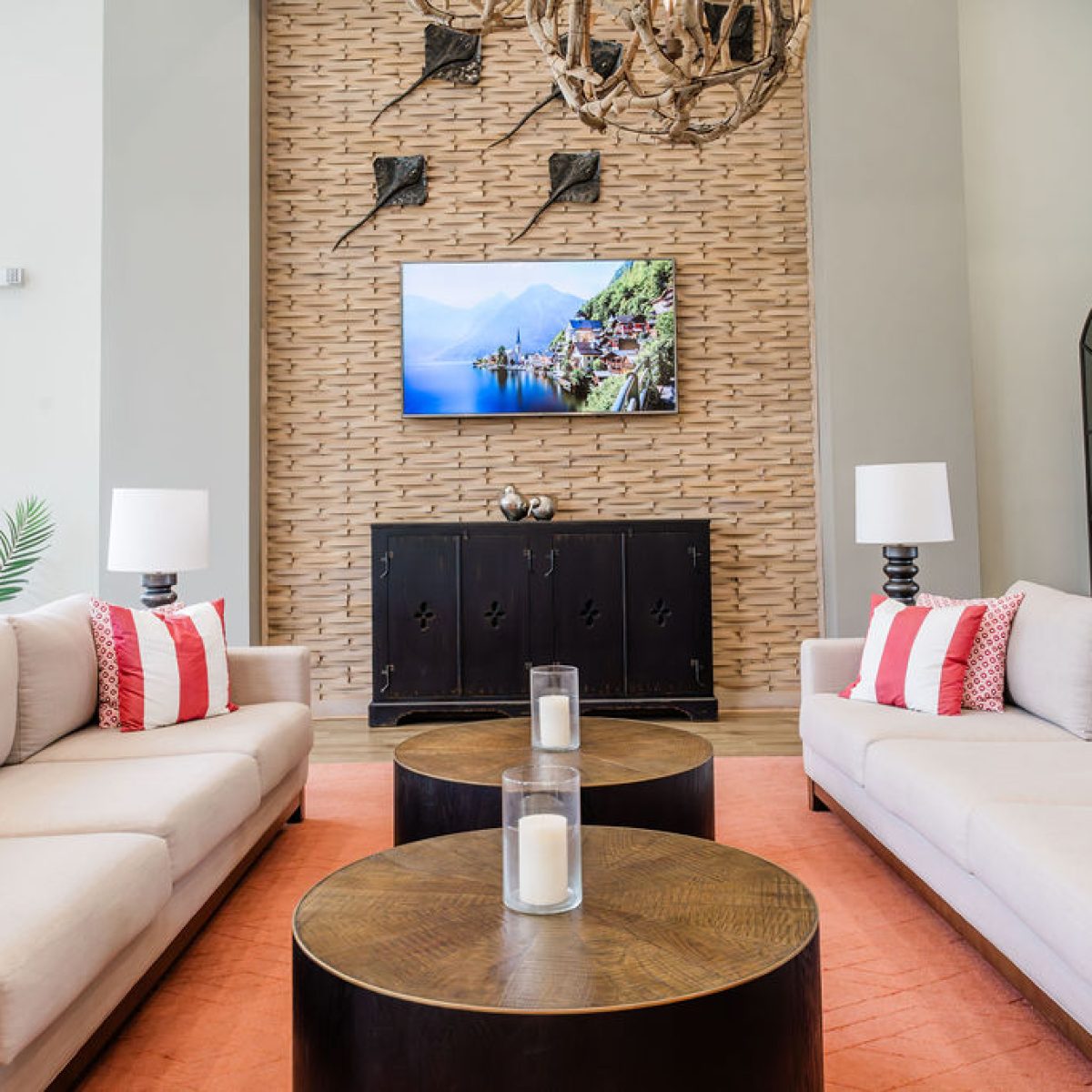 Apartments in Bluffton A modern living room features two beige sofas with red and white pillows, wooden coffee tables with candles, a large wall-mounted TV, and a brick accent wall.