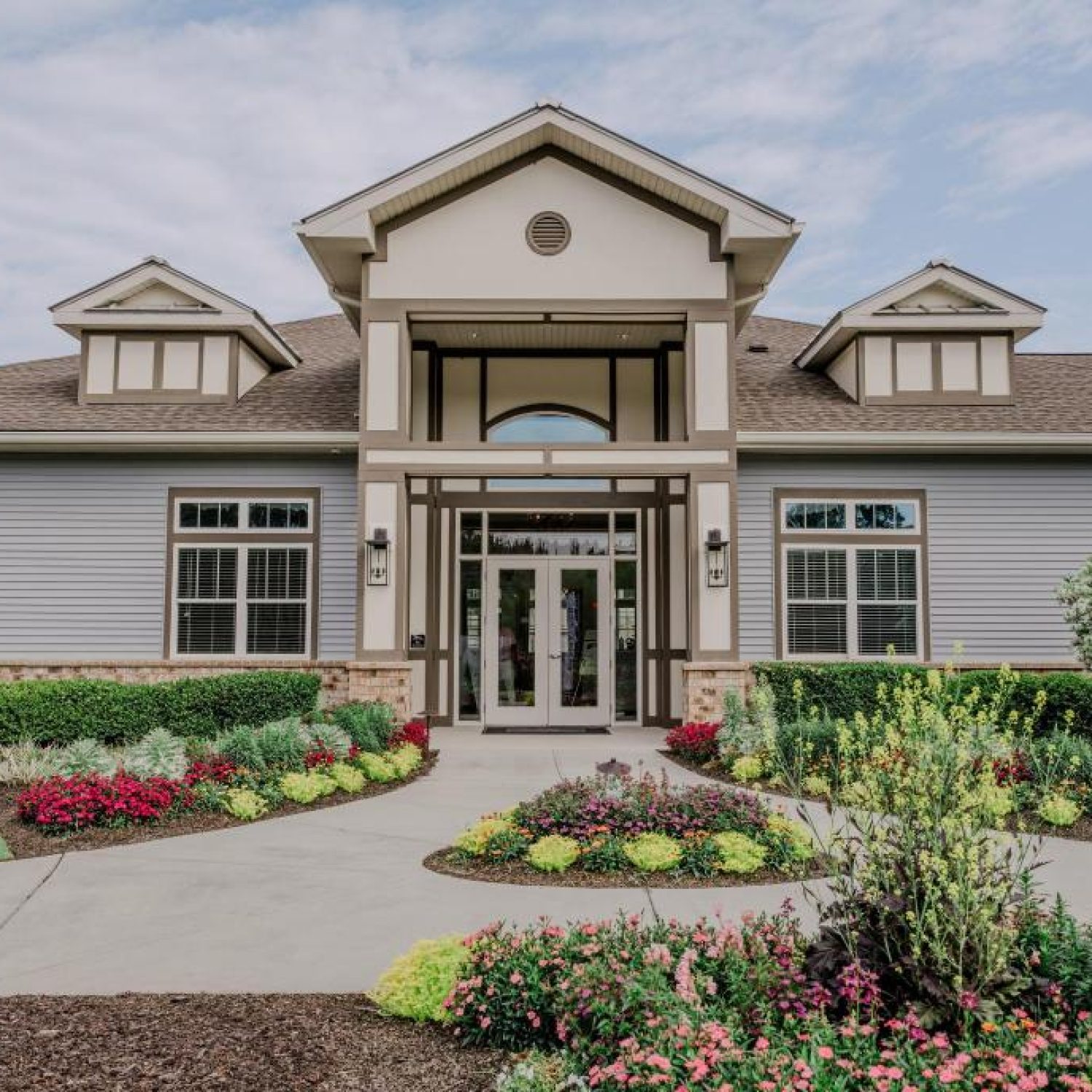 Apartments in Bluffton A modern, single-story building with a symmetrical facade, arched entrance, large windows, and a landscaped garden featuring vibrant flowers and neatly trimmed bushes.