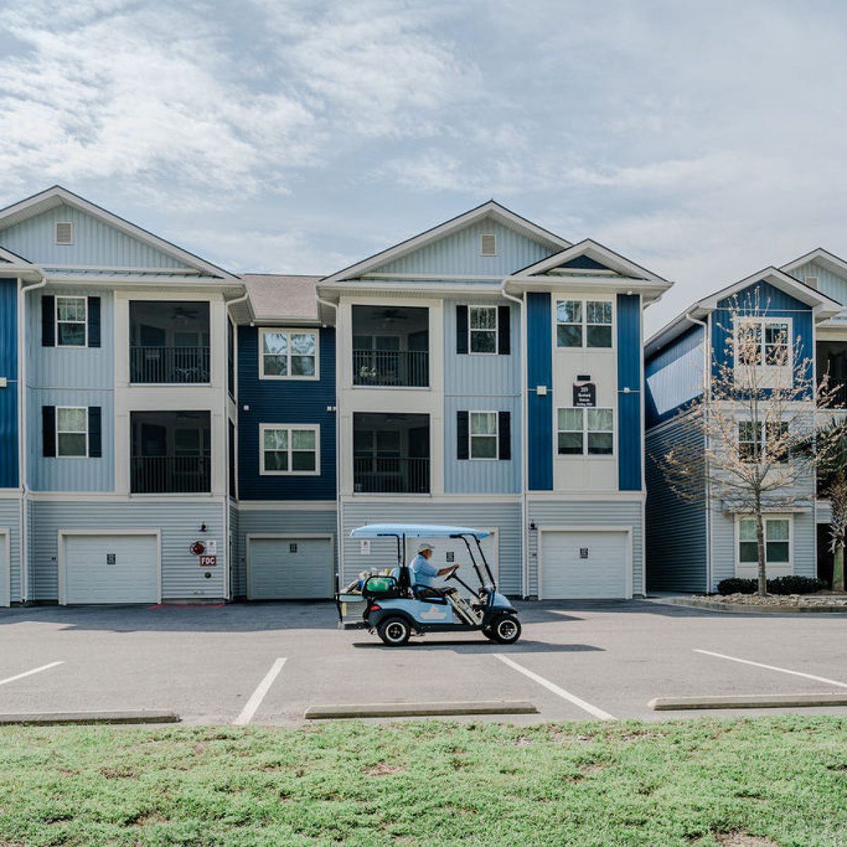 Apartments in Bluffton A three-story apartment building with blue and white exterior features a parking area in the front. A golf cart with one occupant is seen driving in the parking lot.