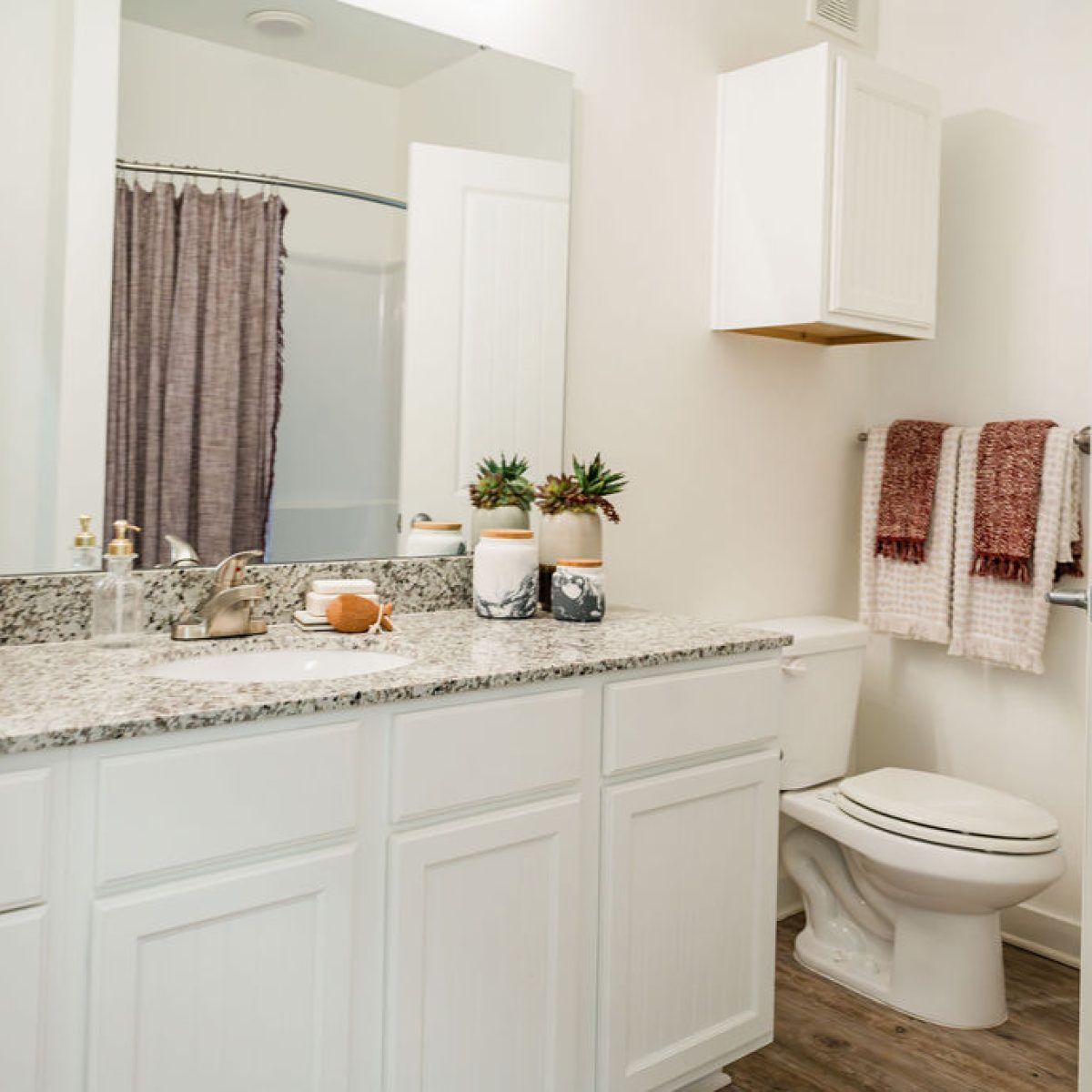 Apartments in Bluffton Modern bathroom with a granite countertop, dual sinks, and white cabinetry. A toilet is next to the counter, with pink and white towels hanging on the wall and potted plants as decor.