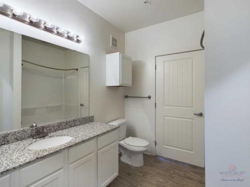 Apartments in Bluffton A bathroom featuring a single vanity with a granite countertop, a wall-mounted cabinet, a toilet, a bathtub shower combo with a curtain rod, and a door. The floor is wood and there's overhead lighting.