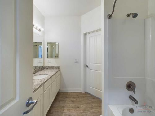 Apartments in Bluffton A bathroom with a granite countertop double sink vanity, a closed door, and a bathtub with a shower curtain rod and wall-mounted showerhead. The floor is wooden.