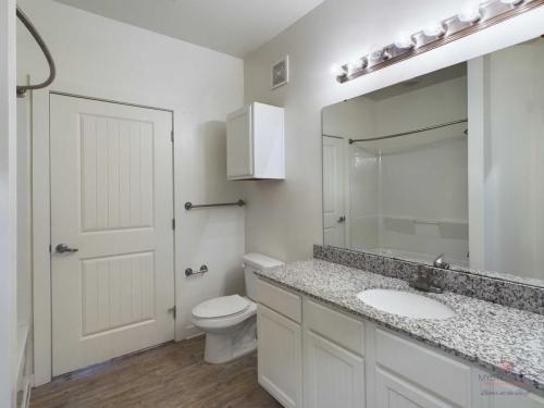 Apartments in Bluffton A bathroom with a granite countertop, mirror with lights, toilet, and shower-tub combination. The walls and cabinets are white, and the floor is wood-patterned.