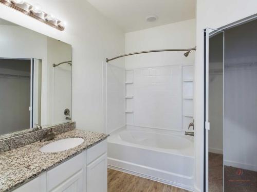 Apartments in Bluffton A bathroom with a granite countertop sink, a mirror with lights above it, a white bathtub with a shower curtain rod, and a built-in shelving unit. The floor is wooden, and there's an open closet door.
