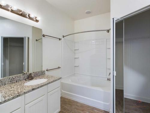 Apartments in Bluffton A bathroom with a granite countertop vanity, a sink, a large mirror with vanity lights, a shower-tub combo with a curved shower rod, and a glass door leading to a walk-in closet.