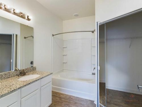 Apartments in Bluffton A bathroom with a granite countertop, white cabinets, a large mirror, and a bathtub with a shower curtain rod installed. The floor is wood, and there's an adjacent closet space.