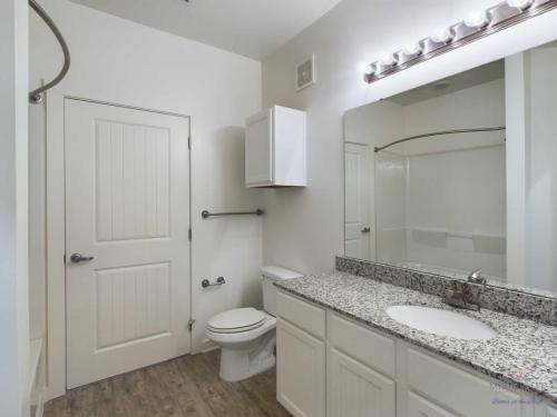 Apartments in Bluffton A bathroom with a white door, toilet, wall cabinet, granite countertop sink, large mirror, and shower in the background. Walls are white, floor is wood-like, and lighting is provided by above-mirror bulbs.