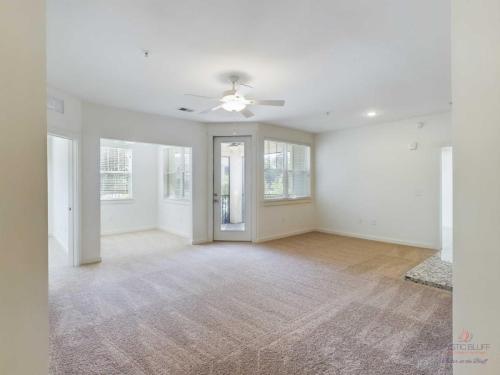 Apartments in Bluffton A bright, empty living room with beige carpet, a ceiling fan, and large windows. A door leads to an outdoor area.