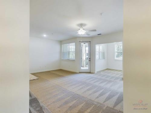 Apartments in Bluffton A bright, empty room with beige carpet, a ceiling fan, large windows, and a door leading to a balcony.