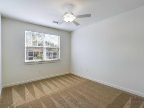 Apartments in Bluffton A bright, empty room with beige carpet, a white ceiling fan, and a window with blinds letting in natural light. The walls are painted white.