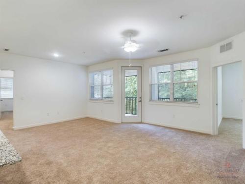 Apartments in Bluffton A bright living room with beige carpet, white walls, a ceiling fan, and multiple windows. There is a door leading to a balcony with a view of trees.