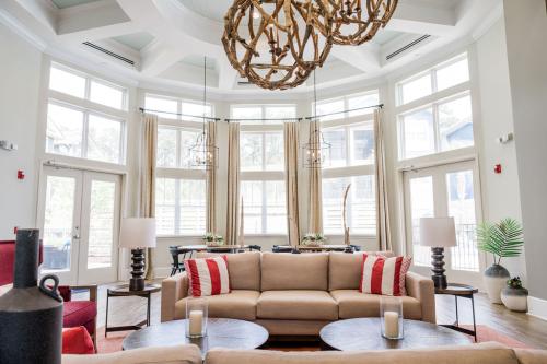 Apartments in Bluffton A bright, spacious living area with large windows, two beige couches, red and white cushions, round coffee tables, and a natural wooden chandelier.
