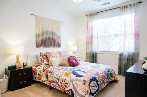 Apartments in Bluffton A brightly lit bedroom featuring a bed with a colorful floral comforter and several cushions, flanked by bedside tables with lamps. A decorative hanging is displayed above the bed, and a large window with sheer curtains.