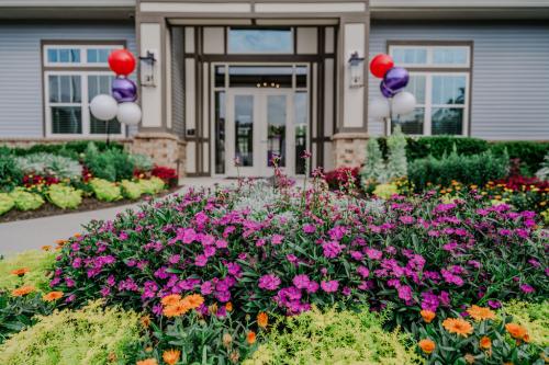 Apartments in Bluffton A building entrance with glass doors is framed by colorful flowerbeds and decorated with red, purple, and white balloons.