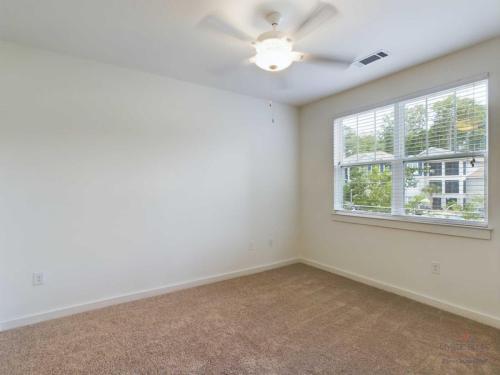 Apartments in Bluffton A carpeted, empty room with white walls, large windows, and a ceiling fan with a light fixture. Natural light enters through the windows, and trees are visible outside.