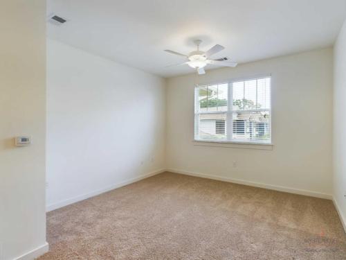 Apartments in Bluffton A clean, empty room with beige carpet, a ceiling fan, and a window with white blinds allowing natural light in.