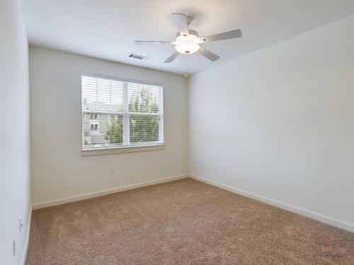 Apartments in Bluffton A clean, empty room with beige carpet, white walls, a ceiling fan with light, and a window with white blinds.