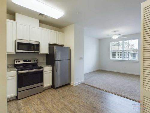 Apartments in Bluffton A compact kitchen with white cabinets, stainless steel appliances, and wood flooring opens to a carpeted living area with a ceiling fan and large window.