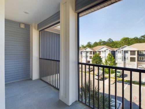 Apartments in Bluffton A covered balcony with gray flooring, white and gray walls, and black railings, offering a view of a residential area with multiple buildings, trees, and parked cars.