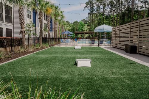 Apartments in Bluffton A fenced outdoor recreation area with artificial grass, two cornhole boards, a wooden privacy wall, and a pool surrounded by patio furniture and umbrellas in the background.