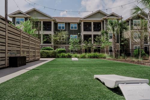 Apartments in Bluffton A green lawn with cornhole boards is in front of a multi-story residential complex with balconies and string lights overhead. A wooden fence lines one side, and several trees and plants are visible.