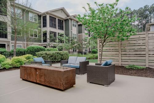 Apartments in Bluffton A landscaped outdoor patio area with wicker seating, blue cushions, a rusted metal fire pit, greenery, and multi-story apartment buildings in the background.