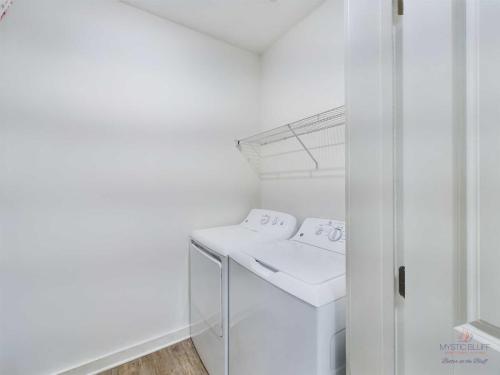 Apartments in Bluffton A laundry room with a washing machine and dryer side by side, under a wire shelf on a white wall, hardwood floor.