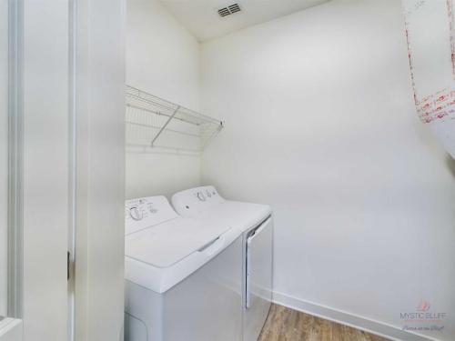 Apartments in Bluffton A laundry room with a white washer and dryer set, a wall-mounted wire shelf above the appliances, and a wooden floor.