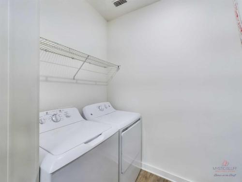 Apartments in Bluffton A laundry room with a white washer and dryer set under a ventilated wire shelf mounted on the wall. The room has white walls and a light wood floor.