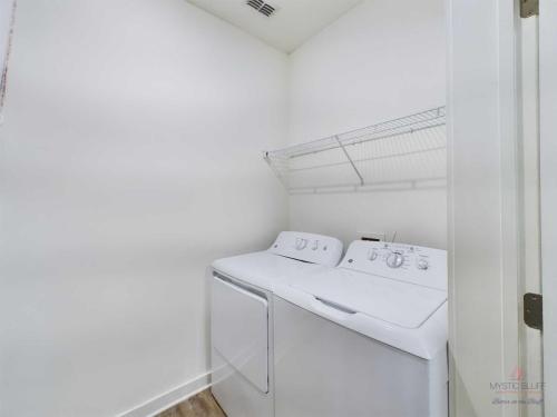 Apartments in Bluffton A laundry room with a white washing machine and dryer side by side, beneath a wire shelf mounted on the white wall. The room has white walls and a wooden floor.