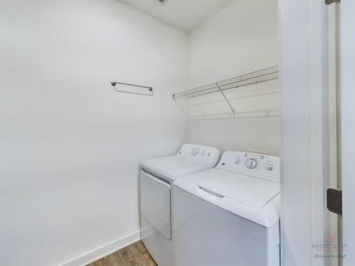 Apartments in Bluffton A laundry room with white washer and dryer units side by side, a metal shelf above them, and a towel bar on the opposite wall. The room has white walls and a wooden floor.