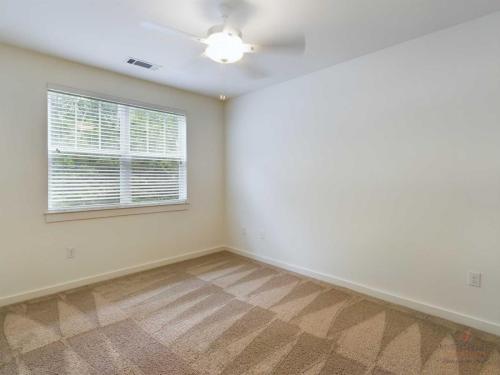 Apartments in Bluffton A minimalistic, empty room with beige carpet, a ceiling fan, and a large window with closed blinds, allowing natural light to enter. Walls are painted white.