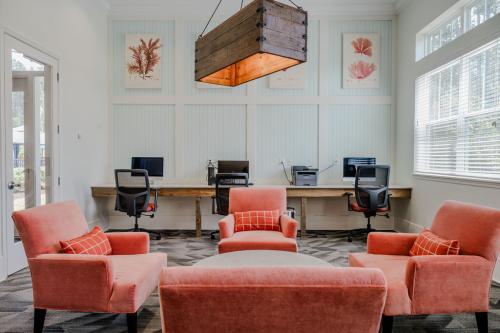 Apartments in Bluffton A modern, airy office space with three desks and chairs along one wall, decorated with coral-themed art. Four salmon-colored armchairs and a round table sit in the center. Large windows provide ample natural light.
