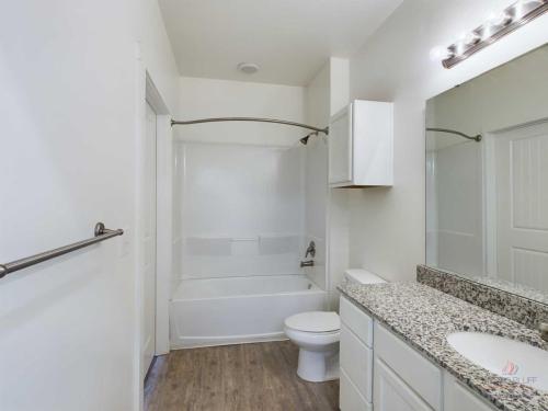 Apartments in Bluffton A modern bathroom with a bathtub, shower curtain, toilet, and a sink with a granite countertop. There is a large mirror above the sink, recessed lighting, and wood-look flooring.