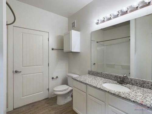 Apartments in Bluffton A modern bathroom with a granite countertop, white cabinets, a toilet, a large mirror with lights, and a shower-tub combo. The bathroom also has a closed door, towel rack, and wood-style flooring.