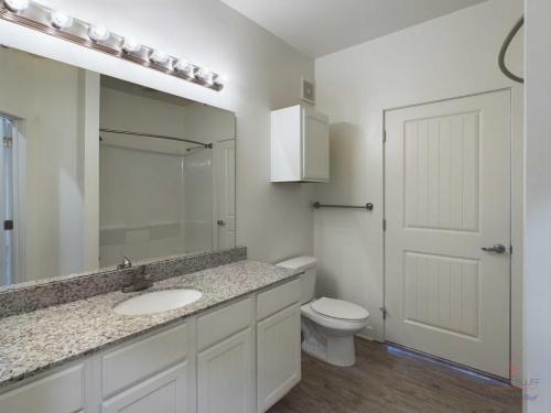 Apartments in Bluffton A modern bathroom with a large mirror, granite countertop, white cabinets, a toilet, and a shower with a curtain. The walls are white, and the floor is wood-patterned.