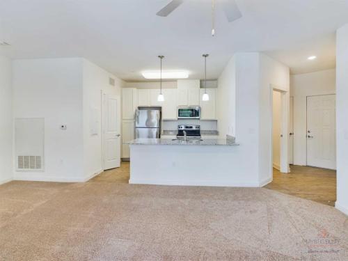 Apartments in Bluffton A modern, empty kitchen and living area with white walls, carpeted flooring, granite countertops, stainless steel appliances, and overhead pendant lights. A ceiling fan is in the center of the living area.