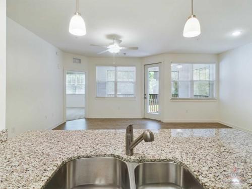 Apartments in Bluffton A modern, empty living area with hardwood floors, white walls, large windows, ceiling fan, and granite countertop kitchen island with stainless steel sink in the foreground.